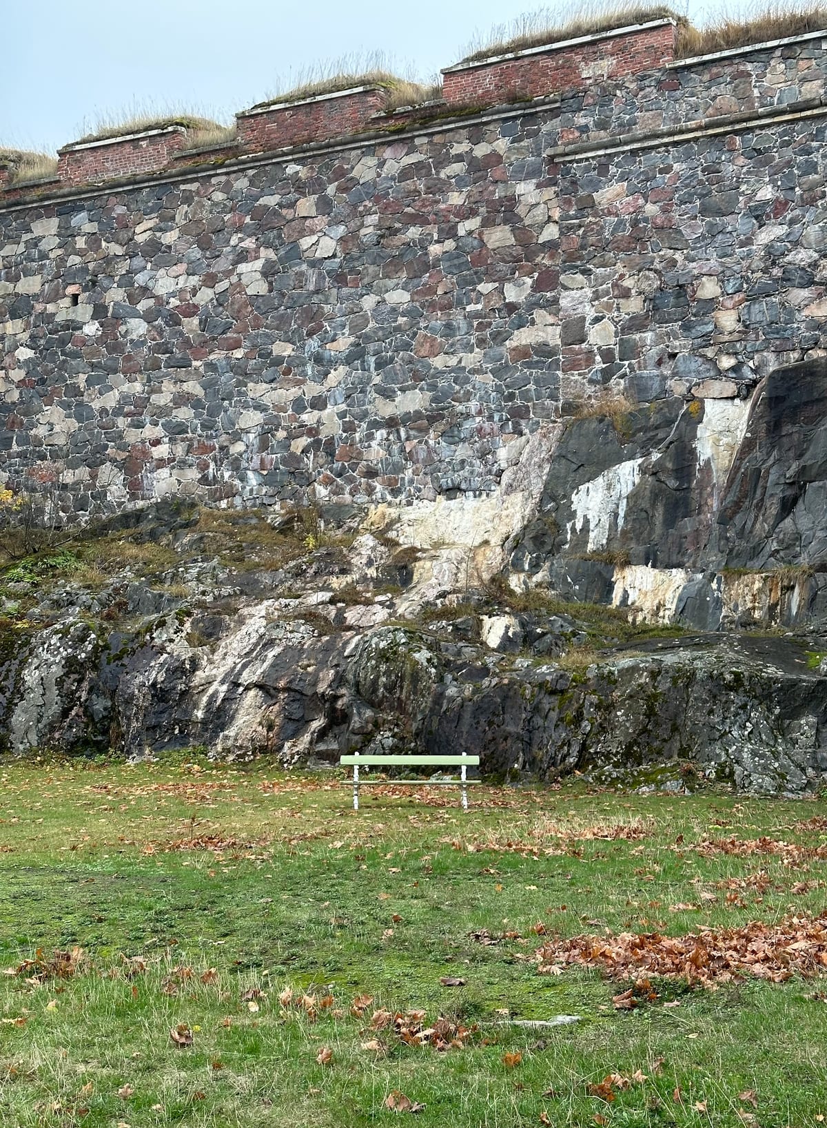 A green bench in front of a large defensive wall made of many gray, dark red and dark blue granite rocks topped off with a brick and mortar gable
